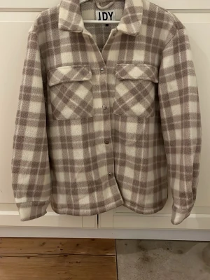Rutig teddyjacka från JDY, M - Mysig teddyjacka från JDY i storlek M. Jackan har ett rutigt mönster i beige och vitt, två bröstfickor med lock och knappar framtill. Perfekt för dig som gillar chill och comfy vibes. Materialet är fluffigt och ger en skön känsla.