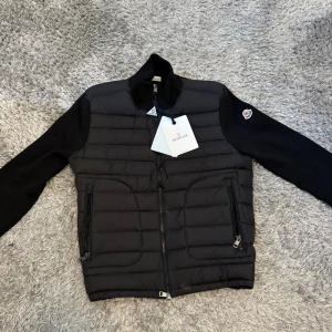 Moncler jacka- herr M  - Helt ny med prislapp kvar. Storlek: M (herr) – modellen är liten i storlek och passar även S. Färg: Svart Äkta vara med certifieringsetikett (DIST och QR-kod för äkthetskontroll). Stilren design med quiltad front och stickade ärmar/rygg,  perfekt för höst/vinter och vår.