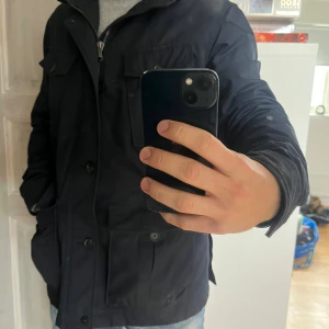 Mörkblå field jacket från Oscar Jacobson - Mörkblå field jacket från Oscar Jacobson med quiltat foder och dragkedja. Jackan har flera stora fickor med knappar, hög krage och luva som går att fälla in/ut (gömma).  Finns även 2 innerfickor 1 med knapp och 1 med dragkedja.