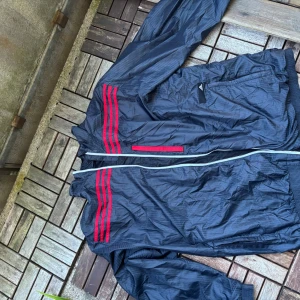 Adidas svart vindjacka med röda ränder - Snygg marinblå vindjacka från Adidas med klassiska röda ränder på axlarna och en röd detalj vid fickan. Jackan har hel dragkedja framtill och ribbade muddar. Perfekt för dig som gillar sportig stil och vill ha något lätt och funktionellt.