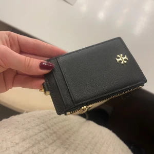 Korthållare Tory Burch - Stilren svart korthållare från Tory Burch i skinn med guldfärgad logga och detaljer.  Det medkommer ett band som man kan fästa på väskan ifall man vill, den går även att ta bort. 🩷