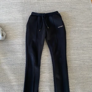 Svarta mjukisbyxor från Les Deux - Svarta mjukisbyxor från Les Deux med snörning i midjan och raka ben. Byxorna har en stilren design med broderad logga på ena sidan och diskreta pressveck framtill. Perfekta för en chill och snygg look.