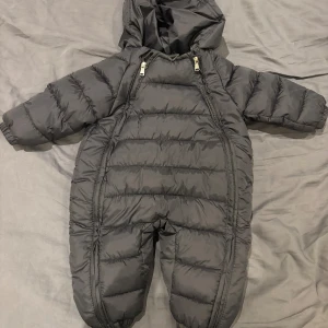 Svart dunoverall med huva, stl 80 - Mysig svart dunoverall med huva och två dragkedjor framtill för enkel påklädning. Overallen är vadderad och har resår vid ärm- och benslut för extra komfort. Perfekt för kalla vinterdagar och håller värmen riktigt bra.