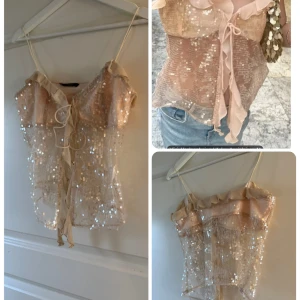 Glittrig beige paljettopp Emmabelin XS-S - Superfin glittrig topp från Emmabelin i beige mesh med paljetter över hela. Tunt material, smala axelband och volangkant upptill. Knytning framtill ger en söt och trendig look. Perfekt att styla med jeans eller kjol för en extra touch.