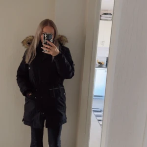 Svart parkas med fuskpäls på huvan - Säljer en svart parkas med stor fluffig huva kantad av brun fuskpäls. Jackan har dragkedja och knappar framtill samt praktiska fickor. Perfekt för kalla dagar med sin rejäla längd och varma material. Snygg och tidlös design som håller dig varm. Väldigt lite använd. 