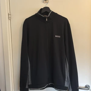 Svart ziptröja från BOSS  - Svart ziptröja från BOSS med grå detaljer på sidorna och vid kragen. Tröjan har hel dragkedja, hög krage och broderad BOSS-logga på bröstet. Materialet är mjukt och stretchigt, perfekt för chill eller sportiga dagar.