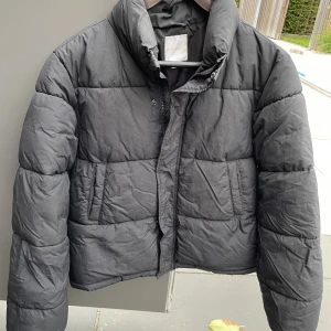 Svart pufferjacka från Gina Tricot - Säljer en svart pufferjacka från Gina Tricot i storlek 158/164. Jackan har hög krage, två sidofickor och stängs med dragkedja och tryckknappar. Perfekt för kalla dagar och har en klassisk, boxig passform.