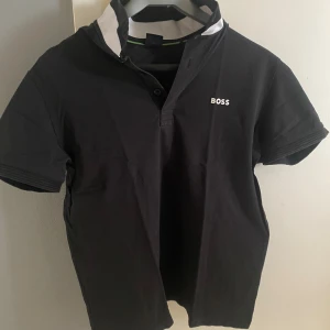 Svart BOSS t-shirt med vit krage - Svart t-shirt från BOSS med vit kontrastkrage och diskret logotyp på bröstet. Klassisk kortärmad modell med knappslå vid halsen. Tillverkad i mjuk bomull som känns skön mot huden. Perfekt för dig som gillar stilrena och enkla plagg med snygga detaljer.