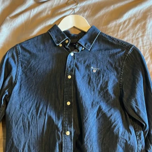 Blå skjorta från GANT - Snygg mörkblå skjorta från GANT med klassisk krage och vita knappar framtill. Skjortan har lång ärm och ett litet broderat GANT-emblem på bröstet. Tillverkad i mjuk bomull som känns skön mot huden. Perfekt för dig som gillar stilrena och tidlösa plagg. Perfekt skick 13-14 years