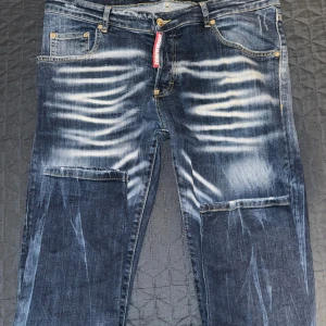 Dsquared2 blå jeans med slitningar - Säljer ett par blå jeans från Dsquared2 med tydliga slitningar och blekta partier framtill. Klassisk femficksmodell med gylf och knapp, samt Dsquared2-logga vid gylfen. Jeansen har en smal passform och är tillverkade i denim med snygg tvätt.