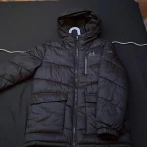Svart pufferjacka från Under Armour - Svart pufferjacka från Under Armour med huva och dragkedja framtill. Jackan har en bröstficka med dragkedja, två stora fickor nertill och  logga på bröstet samt på armen. Perfekt för kalla dagar och riktigt snygg streetstil. Har använts 2 gånger passar även S 