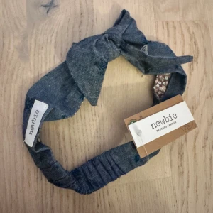Blått diadem med rosett från Newbie pannband  - Snyggt blått diadem från Newbie i bomullstyg med knytdetalj framtill. Utsidan är denimliknande med broderade små blommor och insidan har ett blommigt mönster i lila och vitt. Perfekt accessoar för att lyfta din look. Håraccessoar Ett sött hårband från Newbie med en dekorativ knut. Perfekt för att addera en stilfull detalj till barnets outfit.