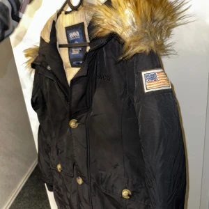 Svart parkasjacka med fuskpäls från Svea - Svart parkasjacka från Svea med beige foder och stor huva med fuskpäls. Jackan har dragkedja och knappar framtill, två stora fickor och en patch med amerikansk flagga på ärmen. Perfekt för kalla dagar och riktigt snygg streetstil.