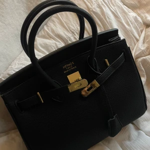 Svart handväska  - svart handväska med guldfärgade metalldetaljer och klassiskt lås. Väskan har två rejäla handtag och en rymlig insida. Perfekt för dig som vill ha en ikonisk och lyxig accessoar med tidlös design.