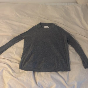 Grå stickad tröja från Lindex - Mysig grå stickad tröja från Holly & whyte med rund hals och ribbade ärmar. Långärmad modell med diskret söm framtill och liten metallbricka vid nederkanten. Perfekt för kyliga dagar och enkel att matcha med jeans eller kjol. Storlek xs/s. 