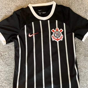 Corinthians svart randig fotbollströja Nike - Säljer en svart fotbollströja från Nike med vita vertikala ränder och Corinthians klubbmärke på bröstet. Tröjan har vit krage och ärmslut samt röd Nike-logga. Tillverkad i Dri-FIT material som andas.