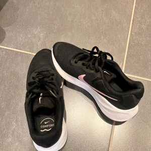 Svarta Nike sneakers  - Snygga svarta Nike sneakers med rosa swoosh och vit sula. Ovandelen är i mesh för bra andningsförmåga och komfort, med klassisk snörning. Sulan har bra grepp och dämpning, perfekt för dig som vill ha både stil och funktion.