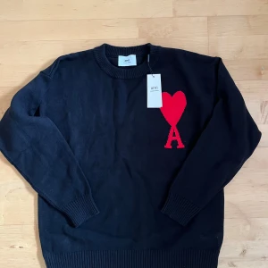 Svart stickad tröja från Ami - Svart stickad tröja från Ami med rund hals och långa ärmar. På bröstet finns ett stort rött hjärta med bokstaven A, vilket ger en unik och cool detalj. Tröjan har ribbade muddar vid ärmslut och nederkant. Perfekt för dig som gillar stilrena plagg med en twist.