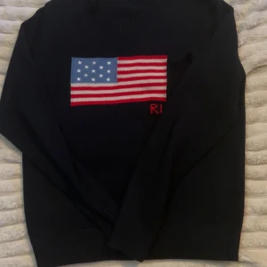 Mörkblå stickad tröja med USA-flagga Ralph lauren - Snygg mörkblå stickad tröja från RL med en stor amerikansk flagga framtill i rött, vitt och blått. Tröjan har rund halsringning, långa ärmar och ribbade muddar. Tillverkad i Italien av merinoull, polyester, akryl och nylon.