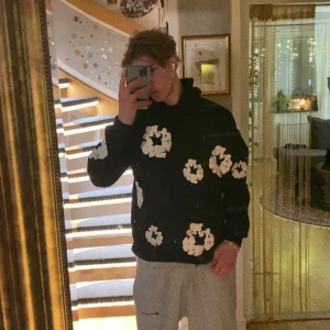 Svart hoodie med vita blommor Denim Tears - Svart hoodie från Denim Tears i storlek L, prydd med stora vita blommor över hela tröjan och huvan. Hoodien har klassisk passform, lång ärm och huva. Perfekt statement-plagg för dig som gillar unika prints och streetwear.