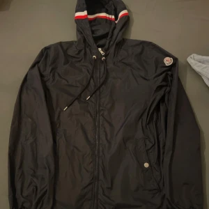 Svart vindjacka från Moncler med huva - Svart vindjacka från Moncler med huva som har röd och vit rand. Jackan har dragkedja framtill, snörning i huvan och en knappstängd ficka. Materialet är glansig polyester och jackan har Moncler-logga på ärmen. Perfekt för dig som gillar stilren och sportig look.