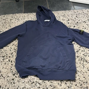 Mörkblå hoodie från Stone Island Junior - Säljer en mörkblå hoodie från Stone Island Junior med klassisk logga på ärmen. Tröjan har huva, känguruficka och är tillverkad i mjuk bomull. Perfekt för dig som gillar streetwear och vill ha något stilrent och bekvämt.