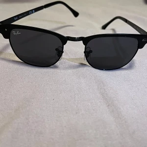 Svarta Ray-Ban Clubmaster solglasögon - Snygga svarta Ray-Ban Clubmaster solglasögon med klassisk halvbåge och mörka linser. Ramen är tillverkad i lätt syntetmaterial och har metall-detaljer. Ikonisk design med Ray-Ban-logga på linsen och insidan av skalmen. Perfekt för dig som gillar stilren och tidlös look.