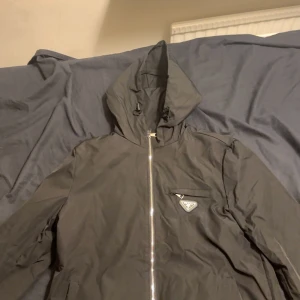 Svart vindjacka med huva från Prada - Svart vindjacka från Prada med ikonisk metallogo på bröstet, hel dragkedja framtill och justerbar huva med snörning. Jackan har en minimalistisk design och är tillverkad i ett lätt syntetmaterial som passar perfekt för blåsiga dagar.