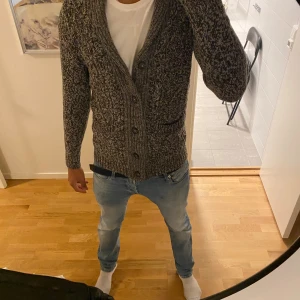 Gråmelerad stickad kofta med knappar - Säljer en gråmelerad stickad kofta med snyggt mönster och stora knappar framtill. Koftan har två fickor och en bred krage som ger en chill och stilren look. Perfekt att slänga på sig över en t-shirt för extra värme.