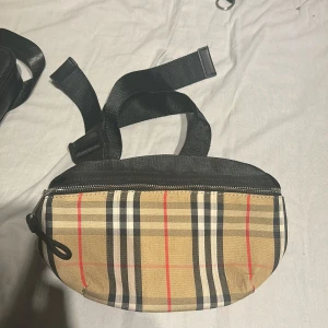 Rutig axelväska från Burberry - Säljer en snygg axelväska från Burberry med klassiskt rutigt mönster i beige, svart, vitt och rött. Väskan har en justerbar svart rem och dragkedja upptill. Perfekt för att bära det viktigaste och ge din outfit en lyxig touch.