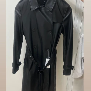 Svart trenchcoat i skinn Mango XS - Säljer en svart trenchcoat/skinnjacka från Mango i skinnimitation. Jackan har klassisk krage, dubbelknäppning, bälte i midjan och spännen vid ärmsluten. Helt oanvänd med prislapp! (Xs men passar S) Pris kan diskuteras☺️