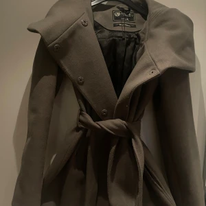 Brun kappa från Amisu Classic Collection - Snygg brun kappa från Amisu Classic Collection med bred krage, knytskärp i midjan och dubbelknäppning framtill. Jackan har långa ärmar och är tillverkad i ett mjukt ull-liknande material. Perfekt för höst och vinter när du vill vara både varm och stilren.