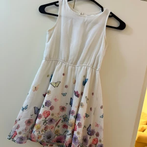 Vit klänning med fjärilar från KappAhl - Superfin vit ärmlös klänning från KappAhl med färgglada fjärilar och blommor nertill. Klänningen har en rund halsringning och ett lätt, luftigt tyg som passar perfekt till varma dagar.