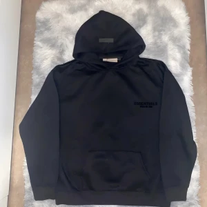 Svart Essentials Fear of God hoodie - Svart hoodie från Essentials Fear of God i storlek L. Klassisk oversized passform med huva och stor känguruficka framtill. Diskret logga på bröstet och stor logga på ryggen. Perfekt för en clean streetwear-look.