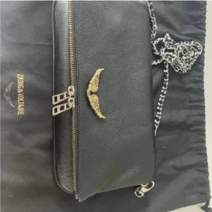 Svart axelväska från Zadig & Voltaire - Svart axelväska i skinn från Zadig & Voltaire med kedjerem i silverfärg och snyggt guldfärgat emblem framtill. Väskan har flera dragkedjor och en stilren, rektangulär form som gör den enkel att matcha med olika outfits. Perfekt storlek för det viktigaste.