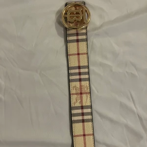 Burberry rutig bälte med guldspänne - Snyggt bälte från Burberry med klassiskt rutigt mönster i beige, svart, rött och vitt. Bältet har ett stort, runt guldspänne med Burberrys ikoniska monogram. Perfekt accessoar för att lyfta din outfit med en lyxig touch.