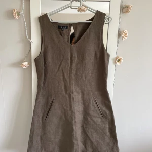 Brun linneklänning från K.U.S Stockholm - Säljer en brun kortklänning i linne från K.U.S Stockholm, storlek 40. Klänningen är ärmlös med v-ringning och har två fickor fram. Baksidan har en snygg knappdetalj och dragkedja. Perfekt för dig som gillar enkel och stilren design.