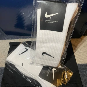Vita Nike strumpor  - Säljer ett par klassiska vita Nike strumpor med svart swoosh-logga. Strumporna är ribbade och har en sportig look som passar perfekt till sneakers. De är tillverkade i mjuk och bekväm bomullsblandning och levereras i originalförpackning.