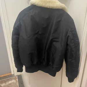 Svart bomberjacka Alpha Industries - Säljer en svart bomberjacka från Alpha Industries med fet vit fuskpäls-krage och röd insida. Jackan har dragkedja, ribbade muddar och fickor på ärmen. Perfekt för dig som vill ha en klassisk och cool look i vinter.