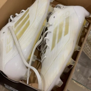 Adidas F50 vita och guld fotbollsskor - Säljer ett par Adidas F50 fotbollsskor i vitt med guldiga detaljer och klassiska tre ränder på sidan. Skorna har snörning, platt sula med dobbar och är tillverkade i syntetmaterial för lätt känsla på planen. Perfekta för dig som vill sticka ut med stil på matchen. Nypris 3000kr. Priset kan diskuteras vid snabb affär
