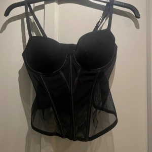 Svart mesh korsett med bygel - Säljer en svart korsett med bygel och vadderade kupor. Toppen har smala axelband och är gjord i meshmaterial med markerade sömmar framtill för en edgy look. Perfekt att styla med jeans eller kjol för en cool outfit. På grund av för stor storlek, så har den dessvärre aldrig kommit till användning.