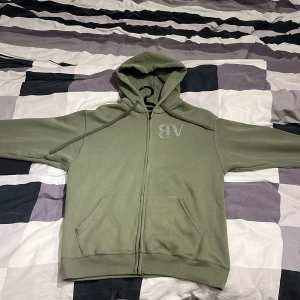 Olivgrön hoodie från Bvaldi med bling - Olivgrön hoodie från Bvaldi med dragkedja och huva. Stora fickor framtill och snyggt blingat BVALDI-tryck både på bröstet och ryggen. Perfekt för dig som gillar streetstyle och vill sticka ut lite extra.