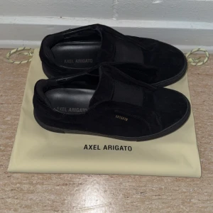 Svarta sneakers från Axel Arigato - Stilrena svarta sneakers från Axel Arigato i mocka med diskret logga i guld på sidan. Skorna har en platt sula och rund tå, perfekt för en clean och minimalistisk look. Kommer med originalpåse.