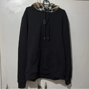 Svart hoodie med rutig huva - Svart hoodie med stor ficka framtill och en snygg rutig huva i beige, svart och brunt. Huva med snörning och klassisk passform. Perfekt för dig som vill ha en stilren men ändå unik look. Tröjan är i storlek xs. Kvitto finns!!