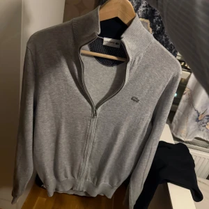 Lacoste kofta  - Snygg grå kofta från Lacoste med hel dragkedja och ribbad krage. Klassisk krokodillogga på bröstet och mjukt material i bomull. 