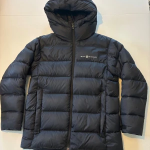 Svart dunjacka från Sail Racing - Svart dunjacka från Sail Racing med huva. Jackan har dragkedja framtill och logga på bröstet. Fylld med 80/20 dun och fjäder, 600 fill power från Allied Feather + Down. Perfekt för kalla dagar och riktigt skön passform.