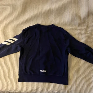 Mörkblå sweatshirt Zadig & Voltaire - Mörkblå sweatshirt från Zadig & Voltaire med vita ränder på ena ärmen och logga nertill på ryggen. Klassisk rund hals och lång ärm, tillverkad i mjuk bomull. Perfekt för en chill och stilren look.