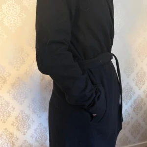 Svart omlottkappa med bälte - Stilren svart kappa i omlottmodell med knytskärp i midjan. Jackan har långa ärmar och en rak passform som ger en clean look. Perfekt för dig som gillar minimalistisk stil och vill ha något som funkar till jeans eller dressat. Köpte på Vinted men passade ej