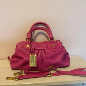 Marc by Marc Jacobs rosa handväska - Snygg rosa handväska från Marc by Marc Jacobs i mjukt skinn med guldfärgade metalldetaljer och avtagbar axelrem. Väskan har dubbla handtag, dekorativa sömmar och en metallbricka framtill. Perfekt storlek för dina essentials och ger en färgklick till din outfit.