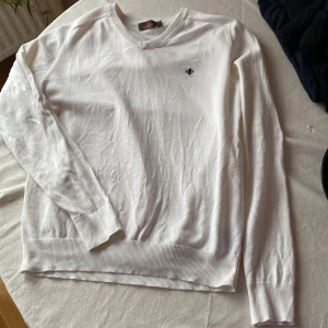 Vit stickad tröja från Morris - Vit stickad tröja från Morris i storlek S. Tröjan har v-ringad hals och ett mörkblått broderat emblem på bröstet. Ribbstickade muddar vid ärmslut och nederkant ger en clean och klassisk look. Perfekt för dig som gillar stilrena plagg.